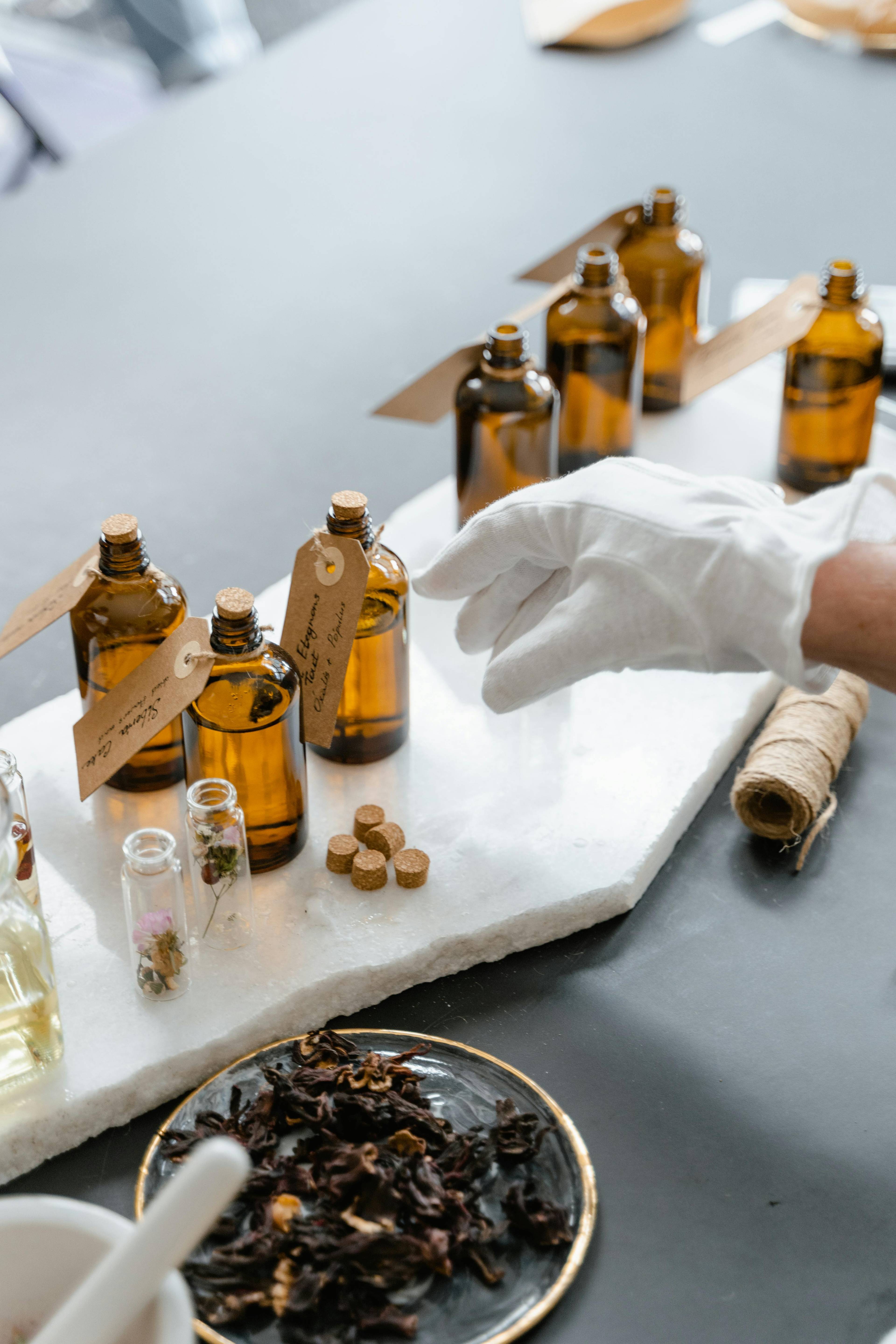 Eine behandschuhte Hand greift nach bernsteinfarbenen Glasflaschen mit Etiketten auf einem Marmortablett, umgeben von kleinen Korken, getrockneten Kräutern und Glasfläschchen.