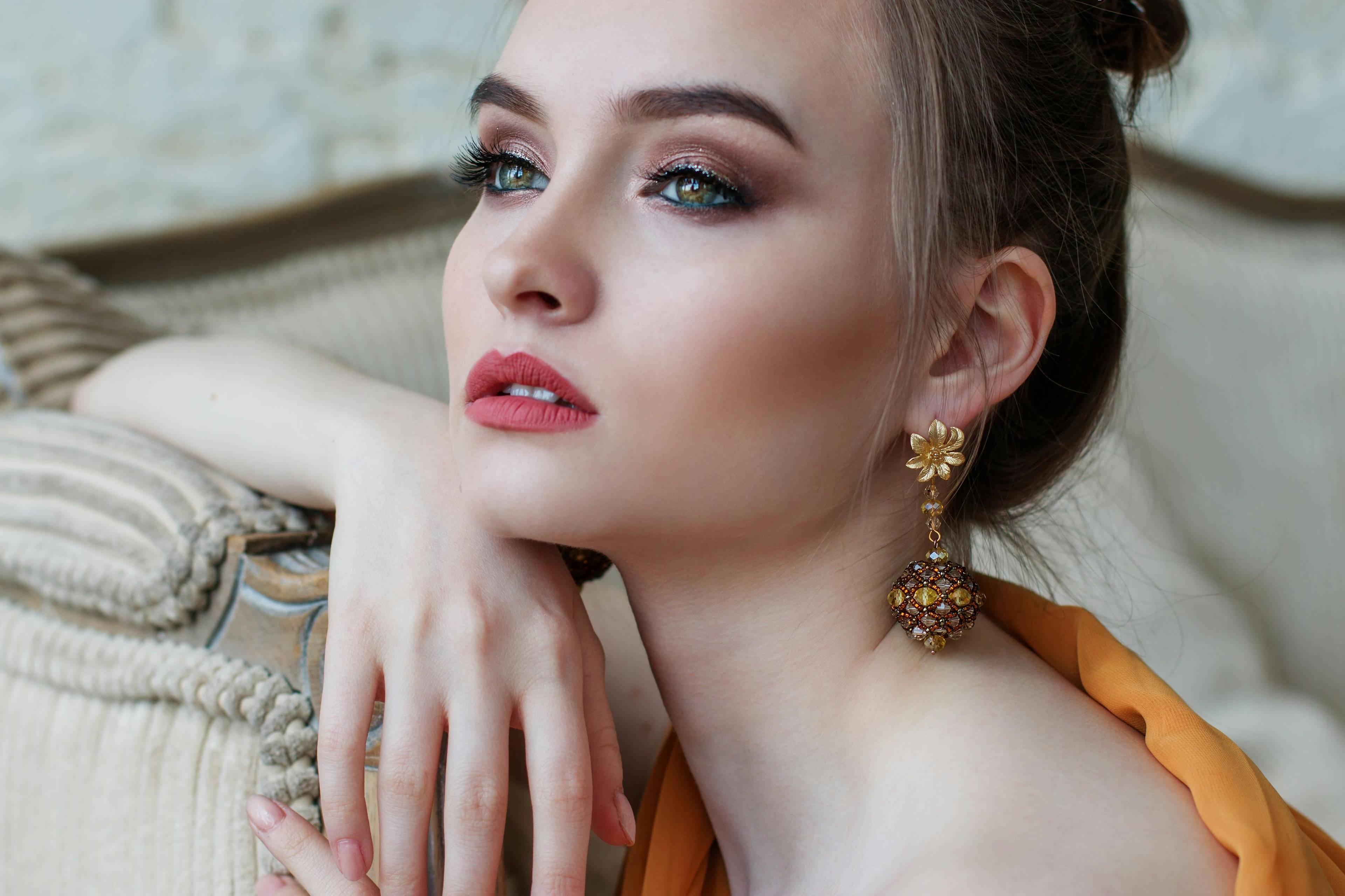 Close-up of a glamorous young woman with glittery eye makeup and floral gold drop earrings, resting her hand on a sofa.