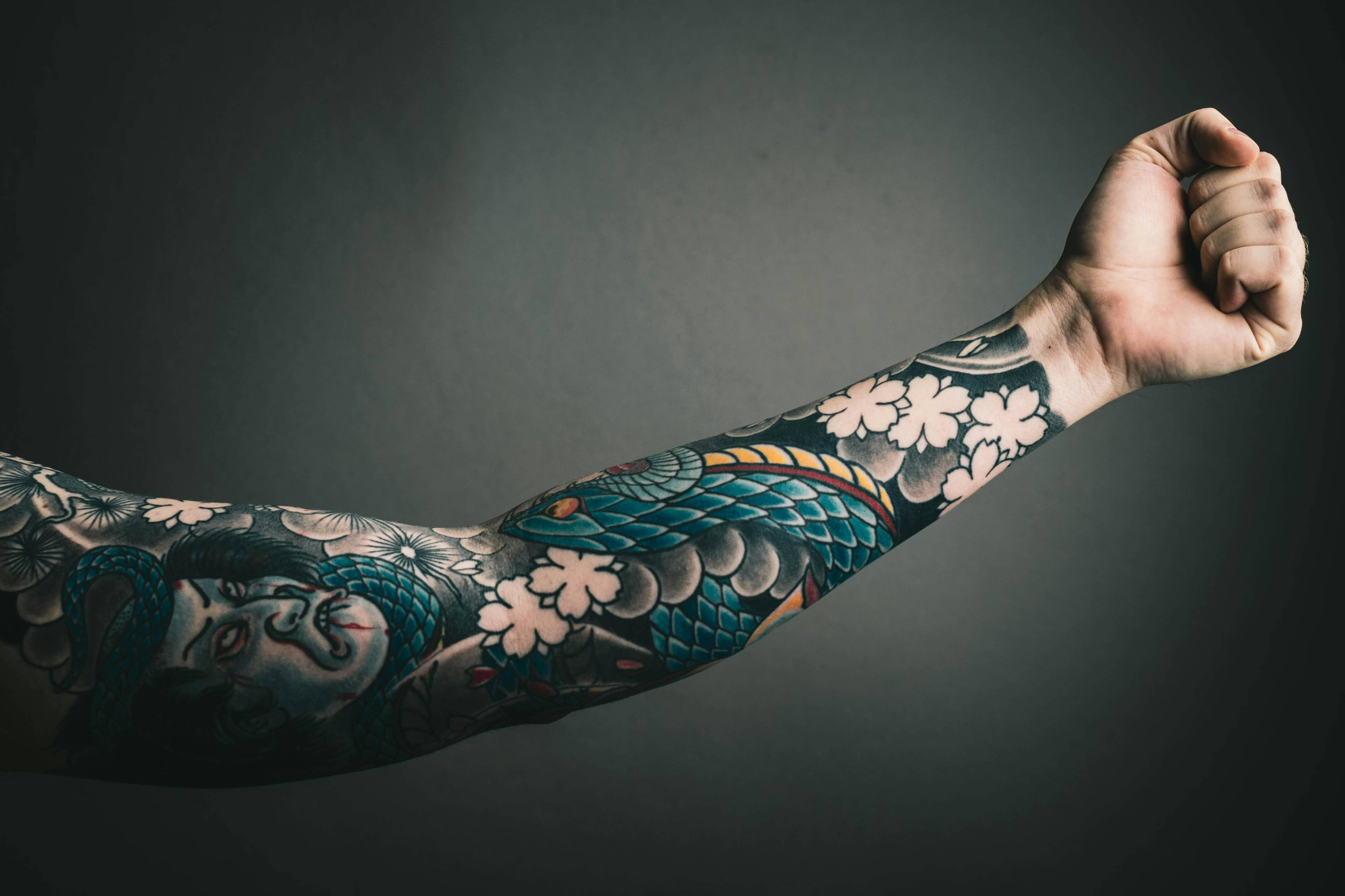 Ein tätowierter Arm mit bunten Motiven, darunter ein Gesicht, Schuppen und Blumen, ist vor einem dunklen Hintergrund zu sehen.
